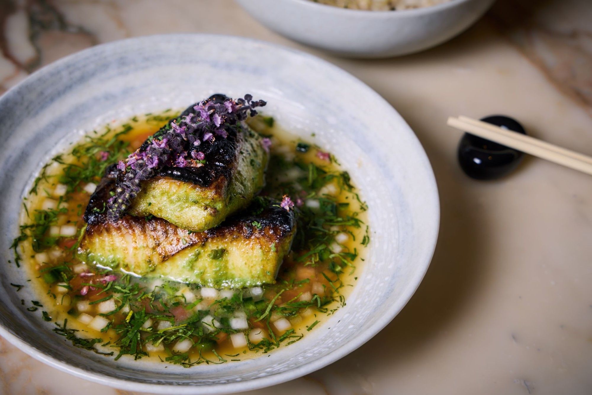 Grilled black cod in a bowl with herb sauce and chopsticks nearby.