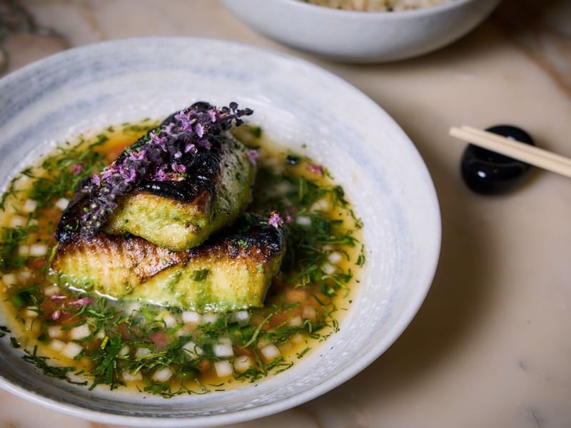 Grilled black cod in a bowl with herb sauce and chopsticks nearby.
