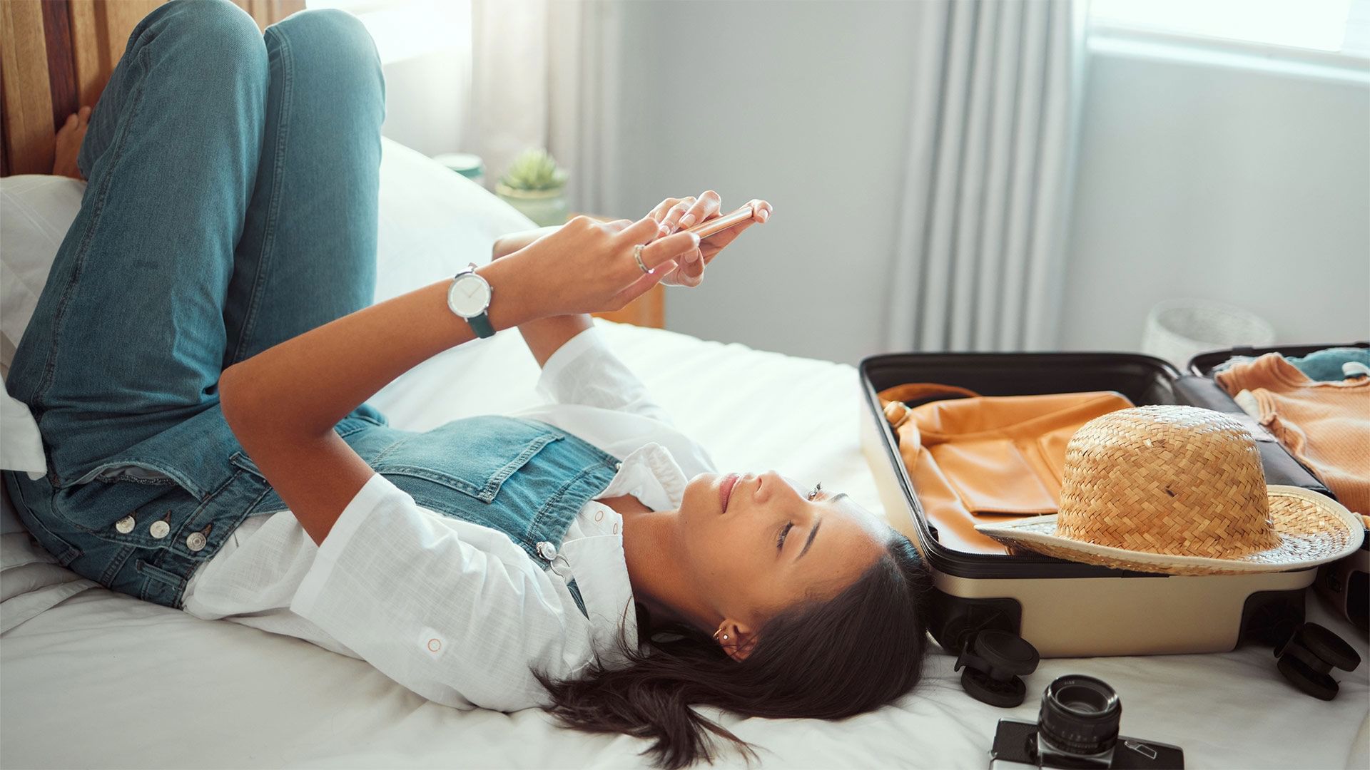 Woman lying on a bed, checking her phone with a suitcase nearby at The Independent