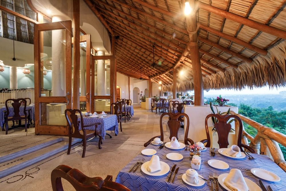 Outdoor dining in Tierra Mia with blue-clothed tables under a large, traditional palapa roof at Quinta Real Acapulco