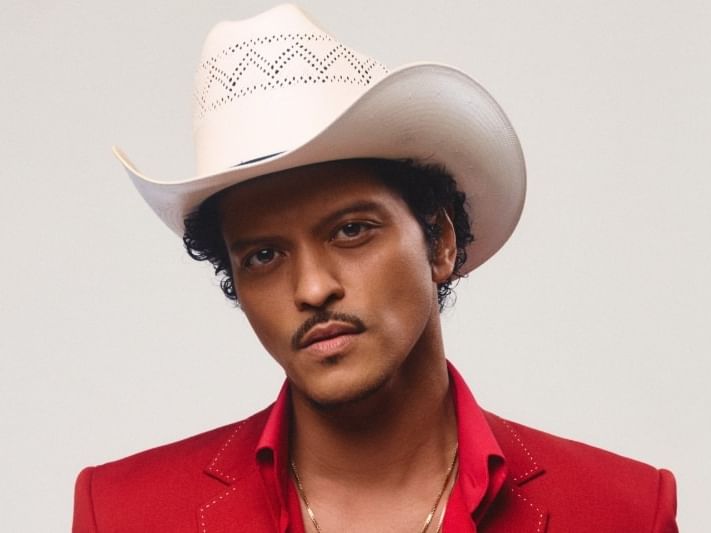 Man wearing white cowboy hat and red embellished suit jacket against neutral background for Bruno Mars The Romantic Tour 2027