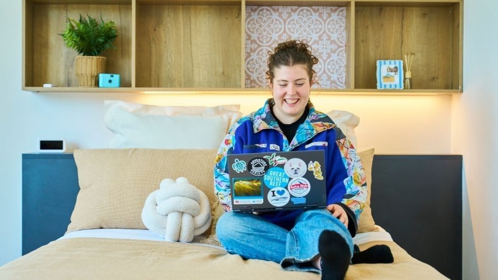 A student smiling and working on a laptop on a bed at UniLodge.