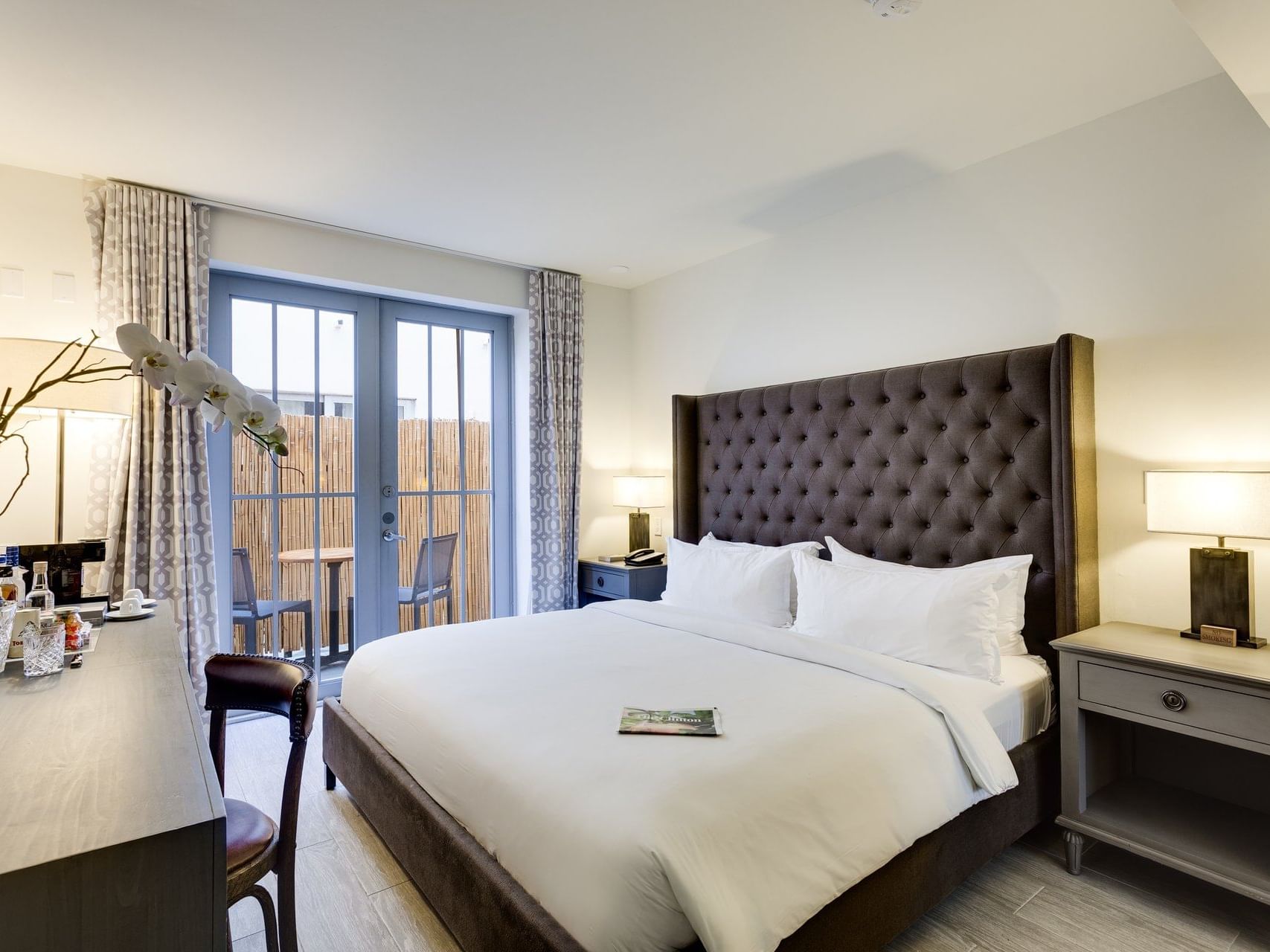 A modern hotel room with a large bed, two nightstands, a desk, and a balcony with outdoor seating.