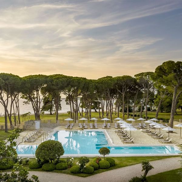 Aerial view of a tranquil resort pool surrounded by trees and sun loungers at dusk in Falkensteiner Hotels & Residences