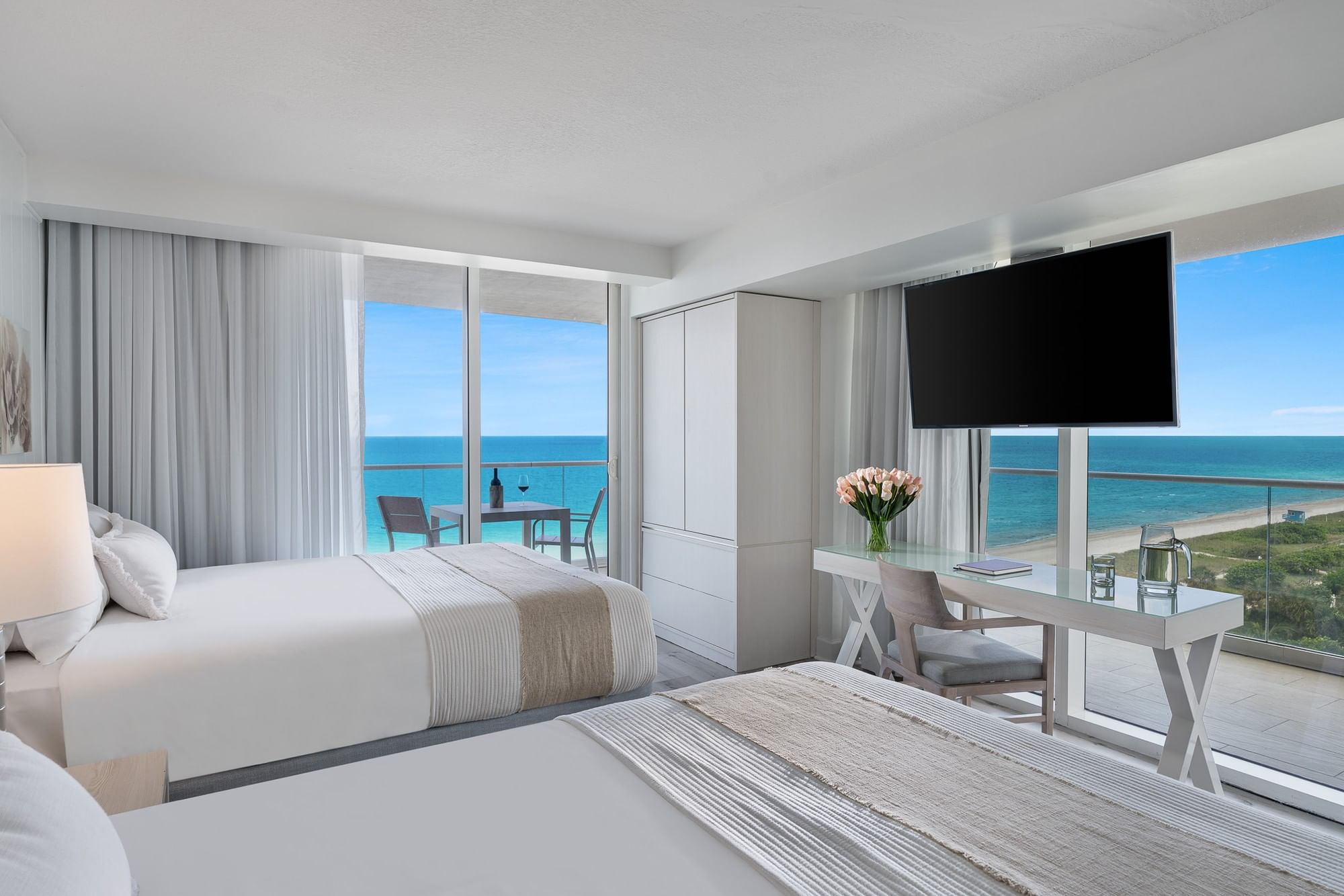 A wide view of a hotel room with two beds, a desk, and a balcony overlooking the ocean.