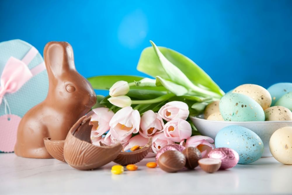 A chocolate bunny and a broken chocolate egg sit beside pink tulips and a bowl of pastel colored Easter eggs. 