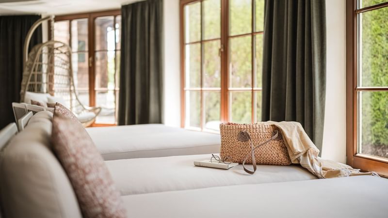 Cozy room with two white beds, a wicker handbag, and large windows with green curtains.