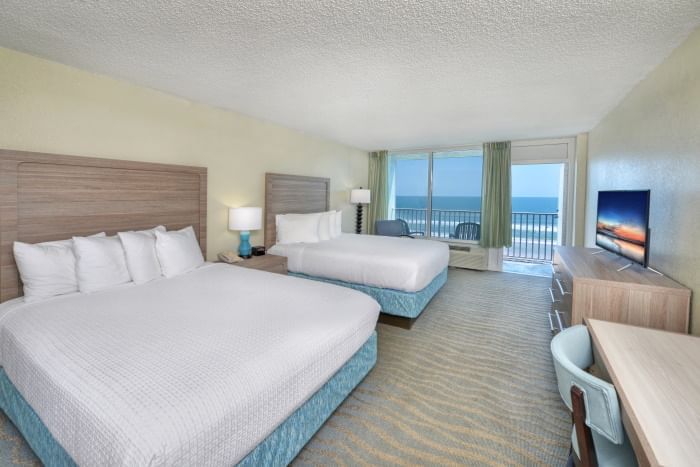 A wide view of a hotel room with two beds, a desk, a TV, and a balcony with an ocean view.