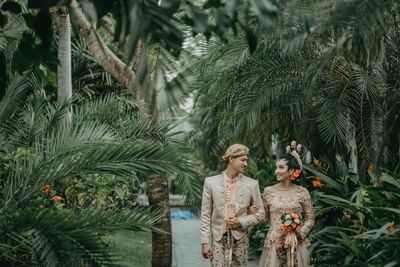 A couple in traditional attire walks hand-in-hand through a tropical garden at The Saujana Hotel Kuala Lumpur