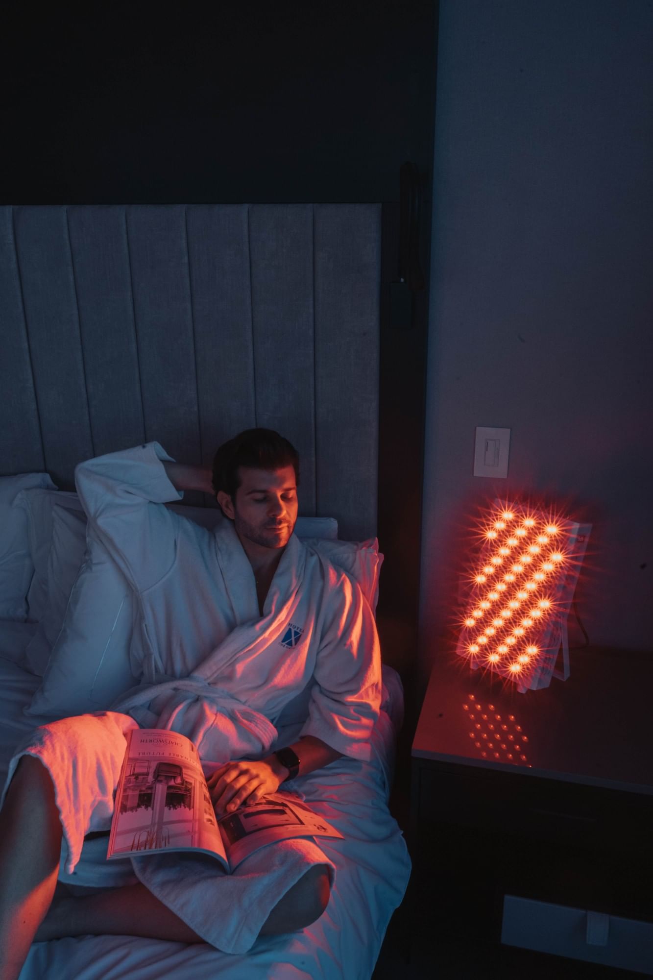 Man in a robe reading by a glowing red light therapy panel on the nightstand at Hotel X Toronto