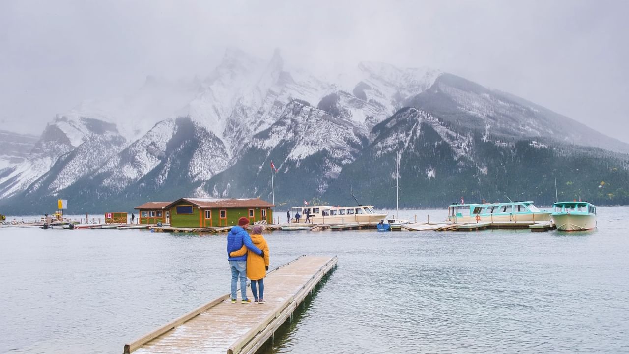 A couple standing on a pier hold each other while admiring a lake and mountain view.