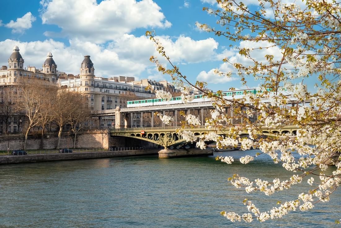 Green train by white blossoms under a blue sky, surrounding a river near Warwick Paris Champs Elysées