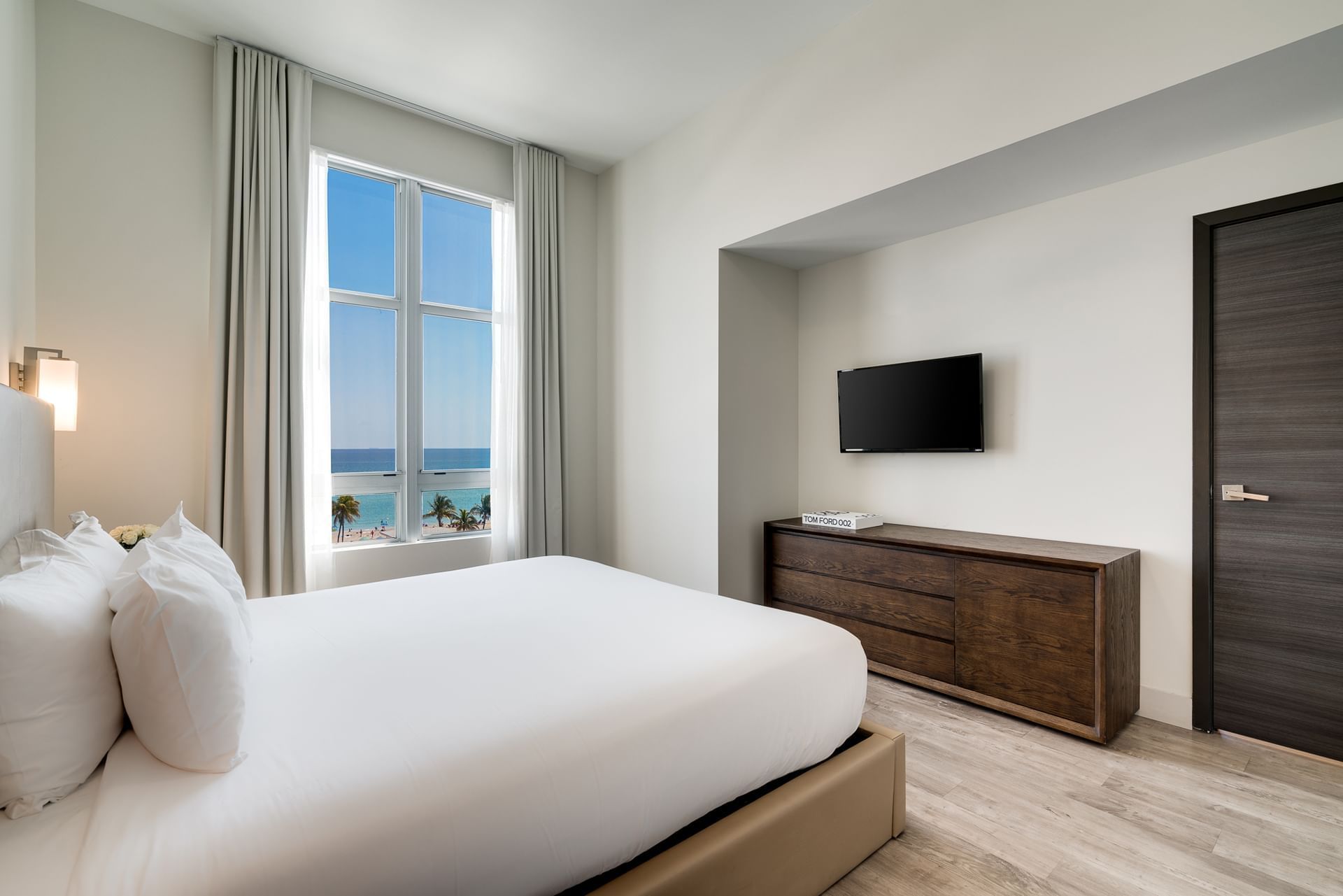 A hotel room with a large window overlooking the ocean, a bed, TV, and dresser.