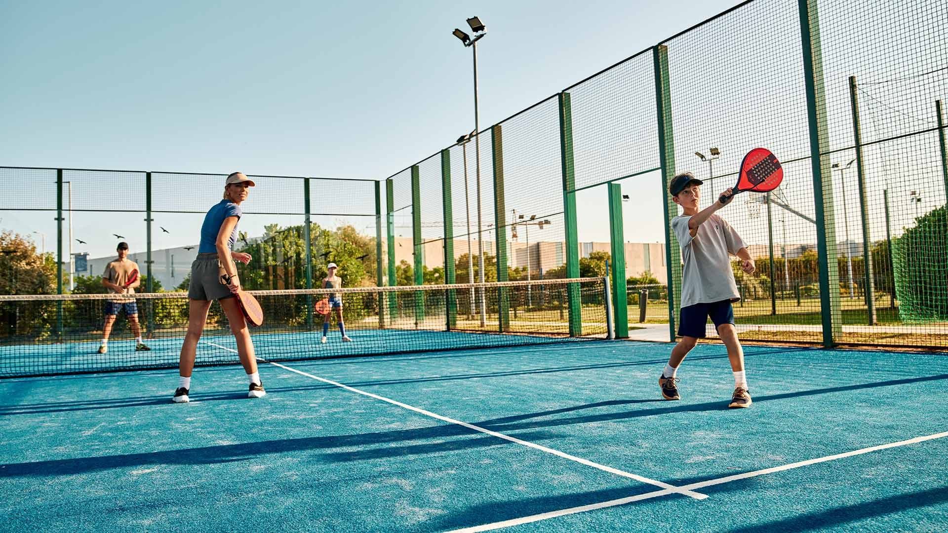 Four people play paddle tennis on a blue court enclosed by a green mesh fence.