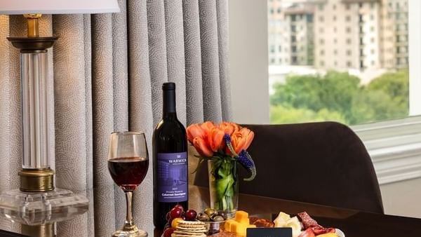Red wine bottle by a cheese platter under a classic lamp with a city view In-Room Dining at Warwick Melrose Dallas