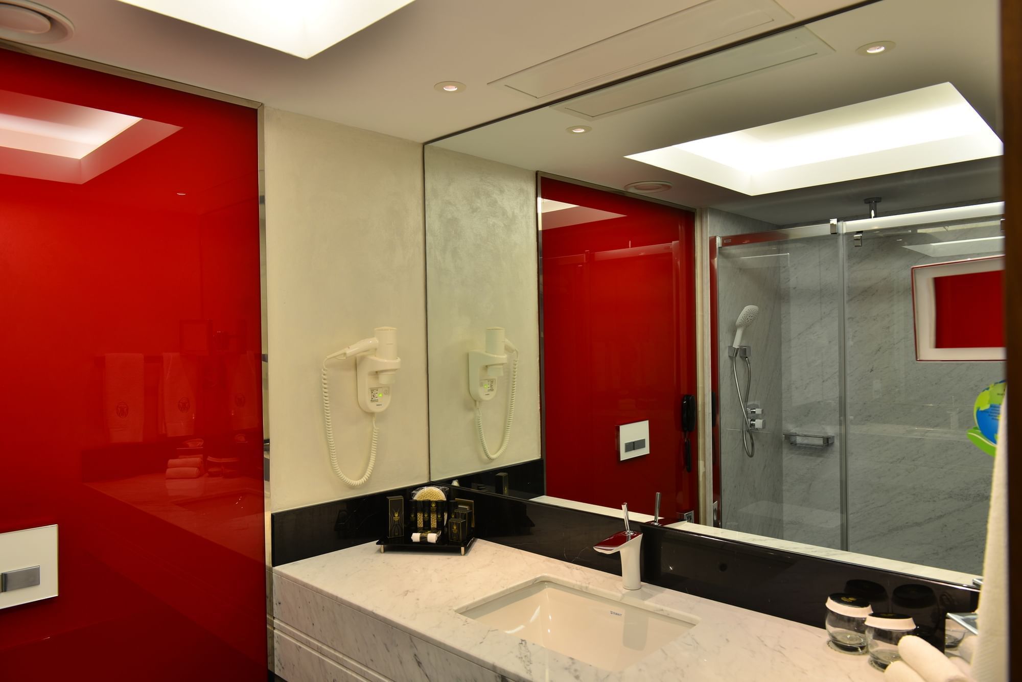 White marble sink by a red glass wall under a large mirror and bright lights in Deluxe Rooms at Warwick Ankara