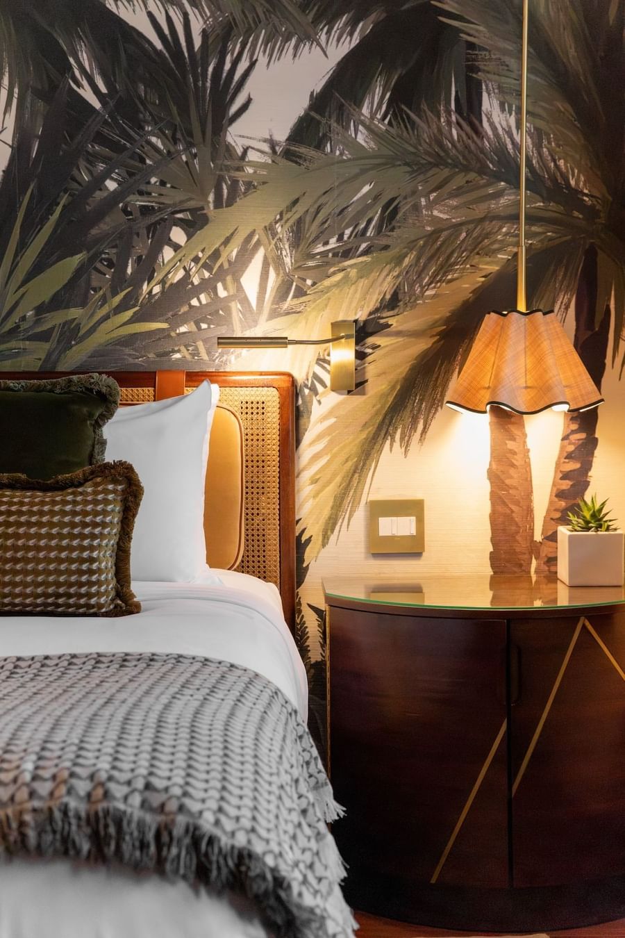 A bed with pillows and a blanket, beside a nightstand with a lamp and plant.