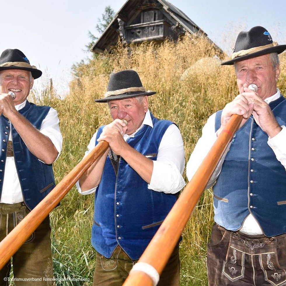 Three men in traditional clothing play wooden horns outdoors in front of a rustic building.