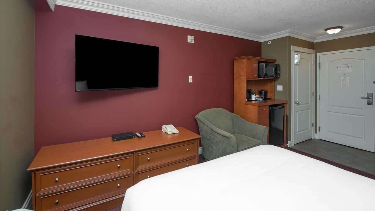 Guest room with burgundy accent wall, flat-screen TV, wooden dresser, and kitchenette at Coast Edson Hotel.