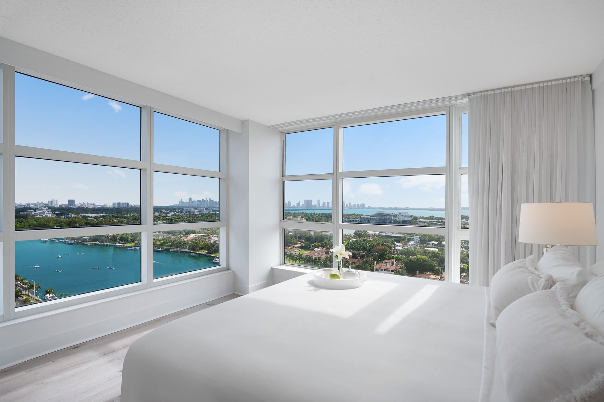 A white bedroom with a king-sized bed and a large window showing a city and water view.