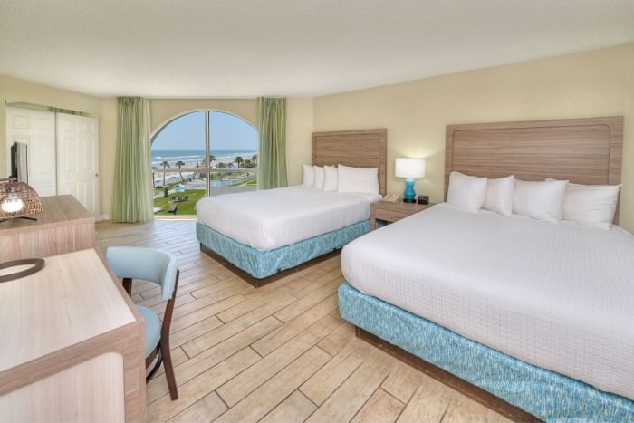 A hotel room with two double beds, a desk, and a chair, with a large window showing a beach view.