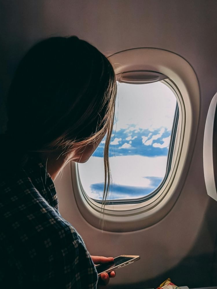 A silhouette of a woman in a plaid shirt holding an iphone looking out an airplane window at clouds.  