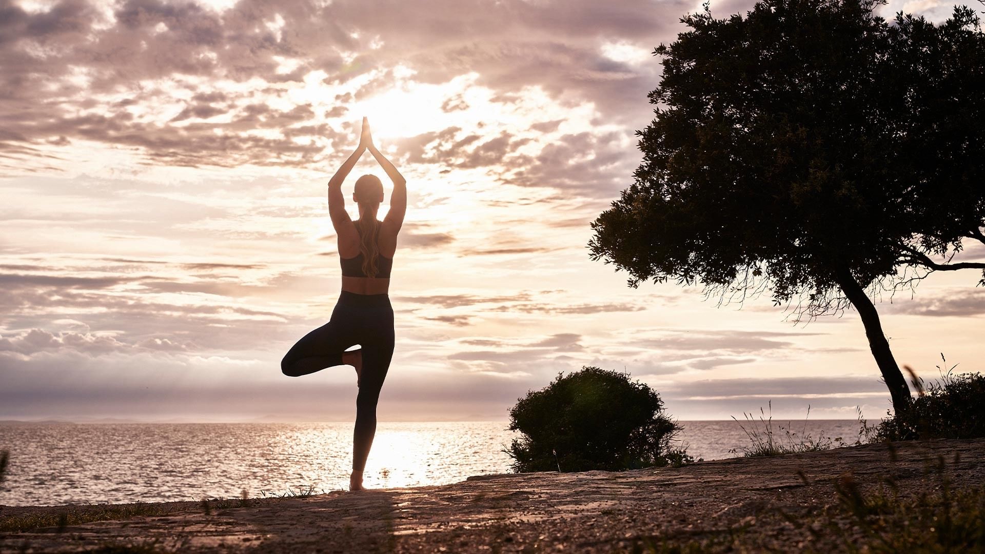 Frau praktiziert Yoga am Meer bei Sonnenuntergang mit Bäumen im Hintergrund.