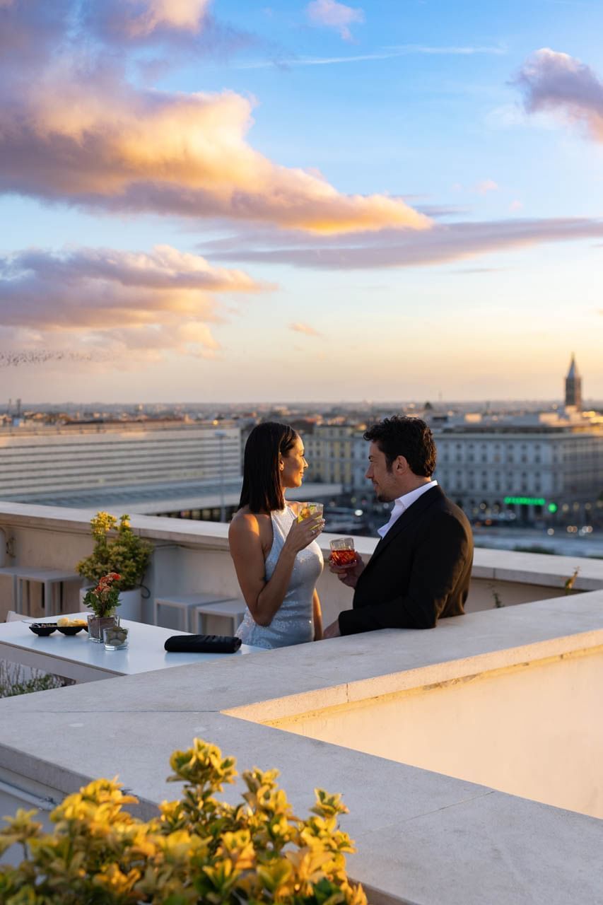 Couple enjoys rooftop drinks at sunset, with a vibrant cityscape and warm, colorful sky at The Independent