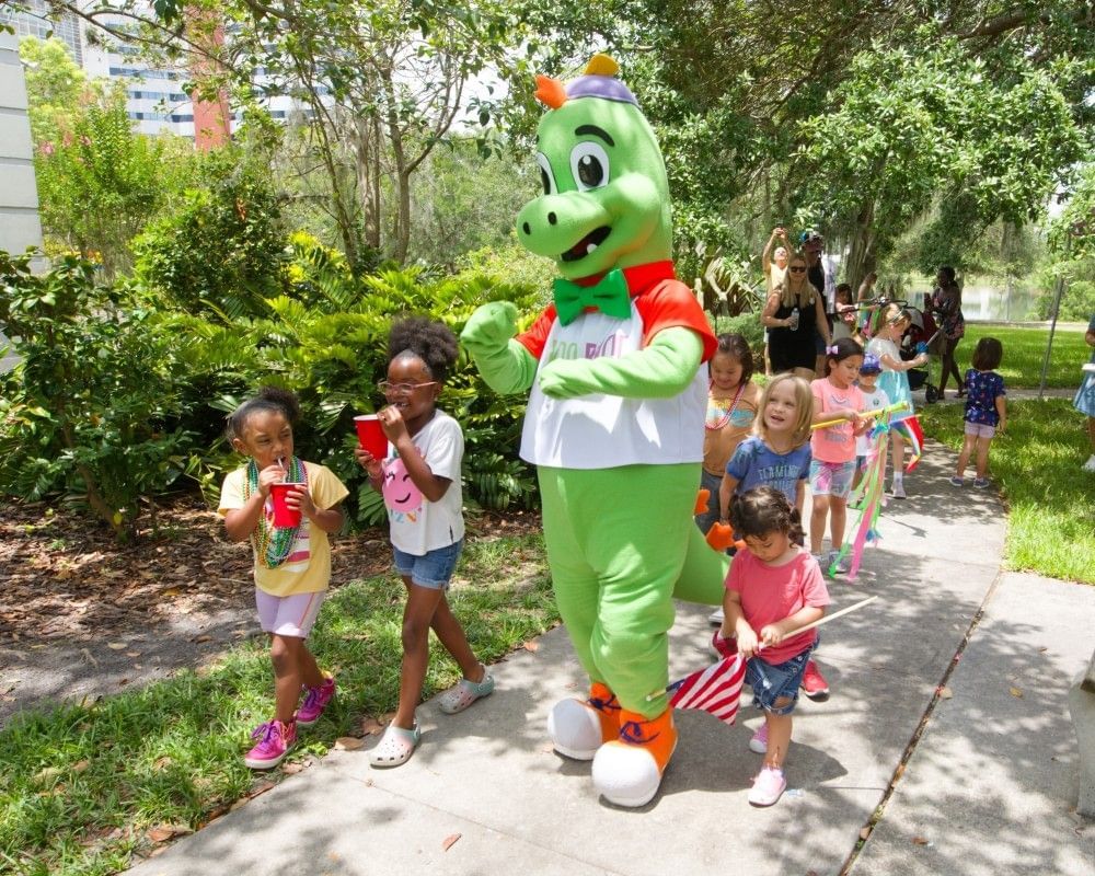 A group of children walk outdoors with a performer in a dinosaur costume.