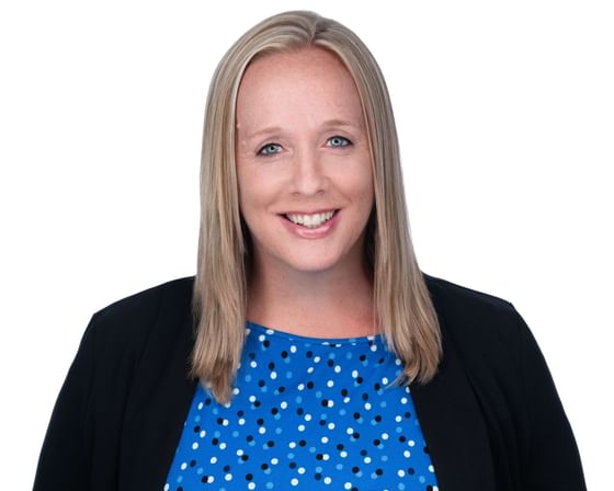Woman with blue eyes, wearing a blue polka-dot shirt and black cardigan, smiling against a white background.