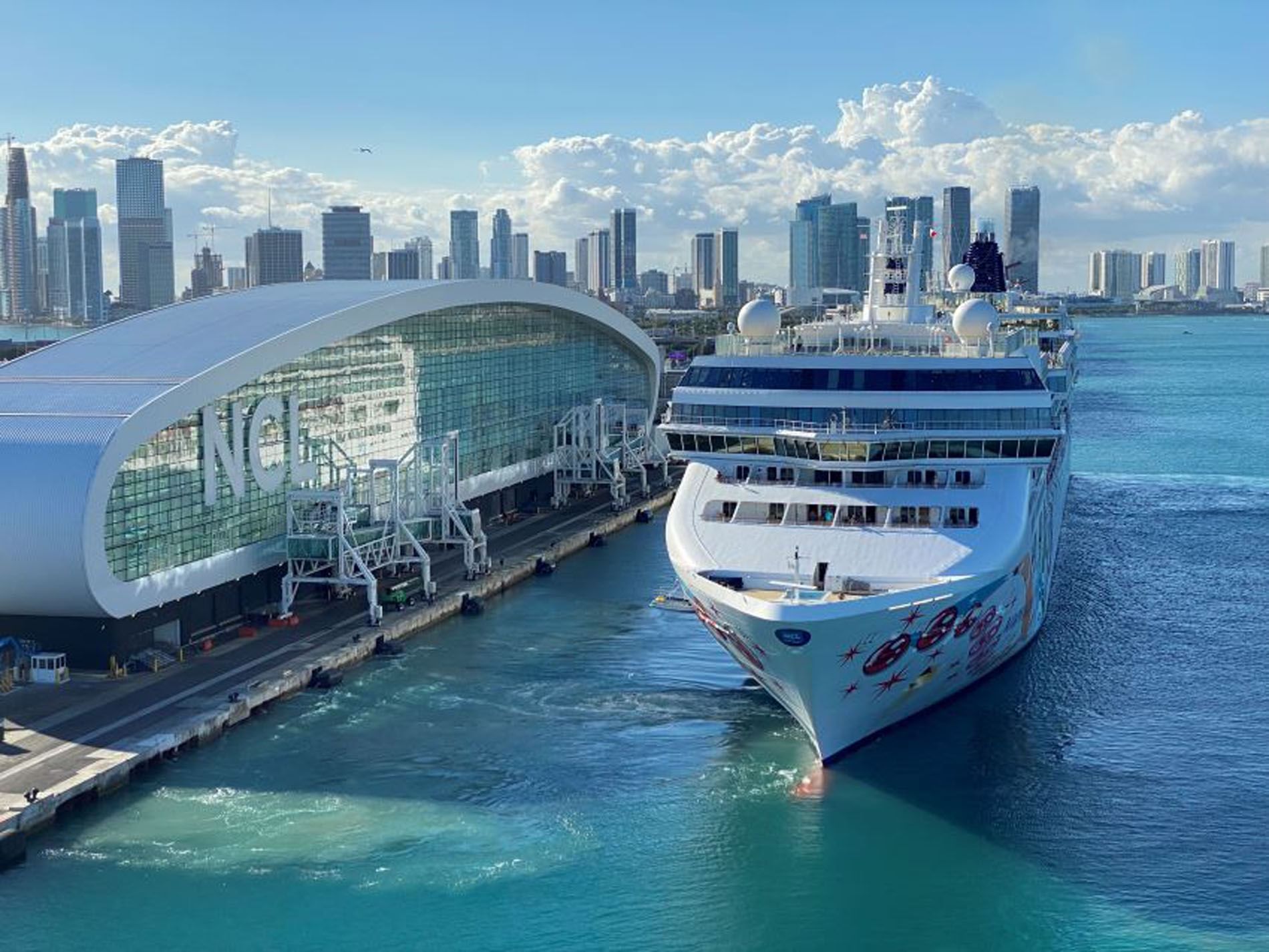 A large cruise ship is docked at a pier with a city skyline in the background.