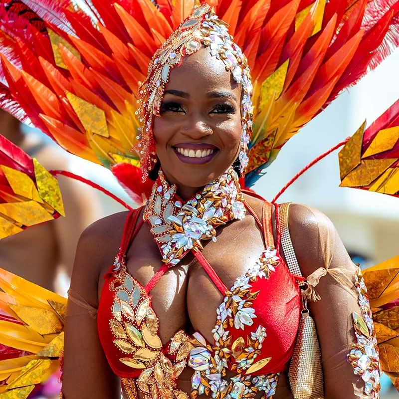 Smiling woman in vibrant red, orange, and yellow feathered carnival costume with jeweled embellishments during Crop Over festival