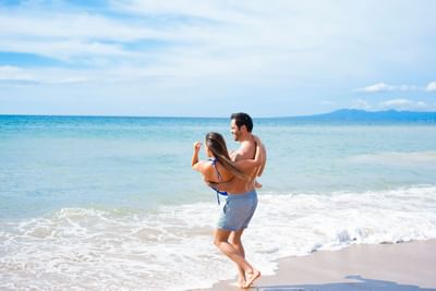 Man holding a woman in his arms while walking through ocean surf on the sunny beach near Casa Dona Susana