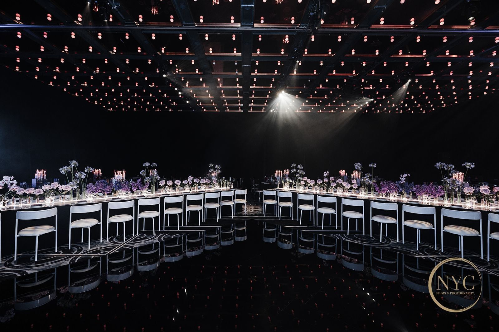 A large banquet hall with two long dining tables, white chairs, floral centerpieces, and a mirrored floor.