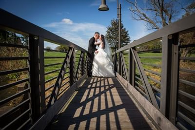 Bride and groom embrace on a wooden bridge with green lawns and blue sky at Aramark Chauncey Conference Center