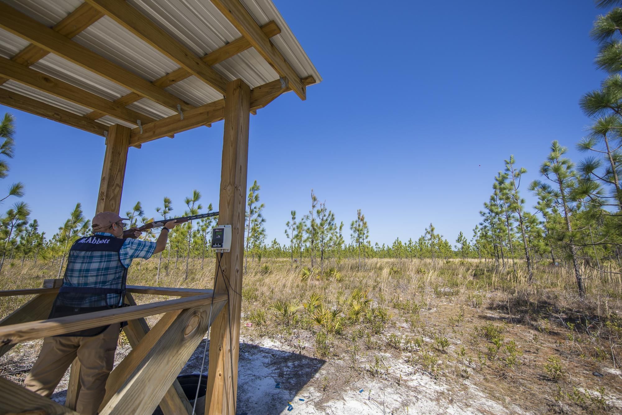 Man with a shotgun on a wooden shooting stand aiming at a clay target in a field.
