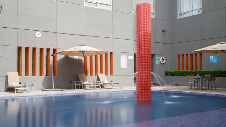 Refreshing outdoor pool featuring a red fountain pillar, lounge chairs, and sun umbrellas at Real Inn Torreon