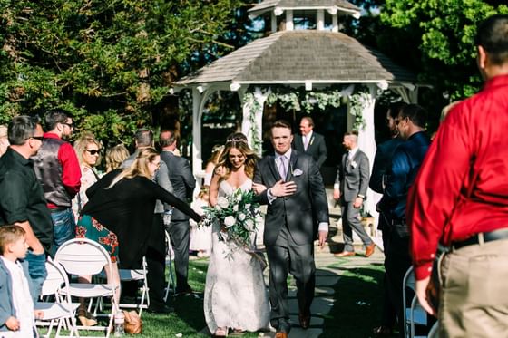 Newlywed couple walking down the aisle after outdoor ceremony with flower-decorated gazebo at Cambria Pines Lodge