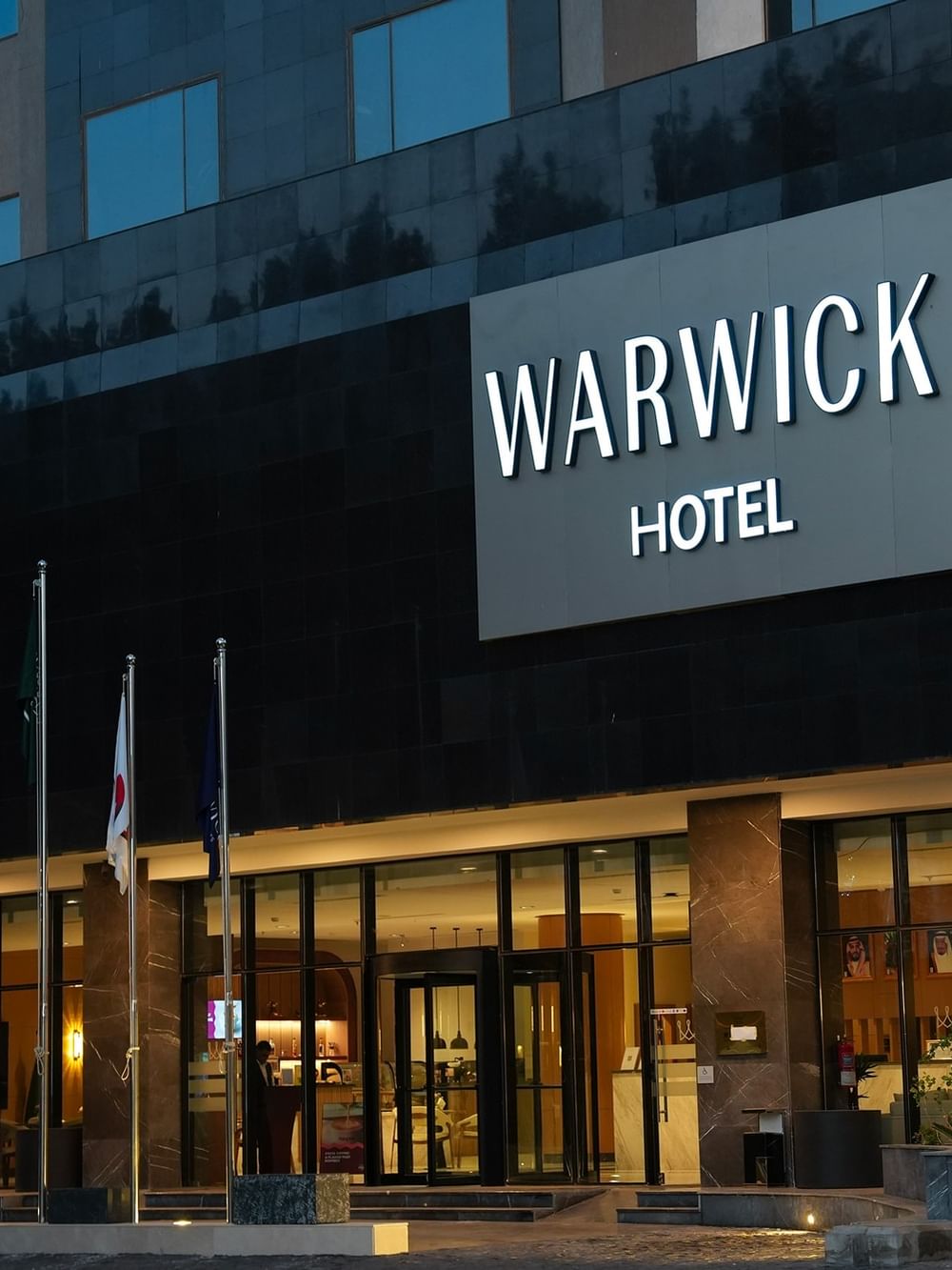 Illuminated Warwick Hotel entrance at night with flags and a revolving door.