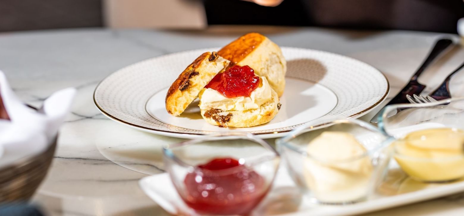 Close-up of a scone with jam and cream, and hands serving a teacup for afternoon tea at The Londoner Hotel