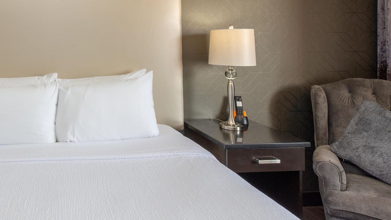 Guest room with white bedding, nightstand with lamp, and upholstered chair at Coast Calgary University Area Hotel by APA