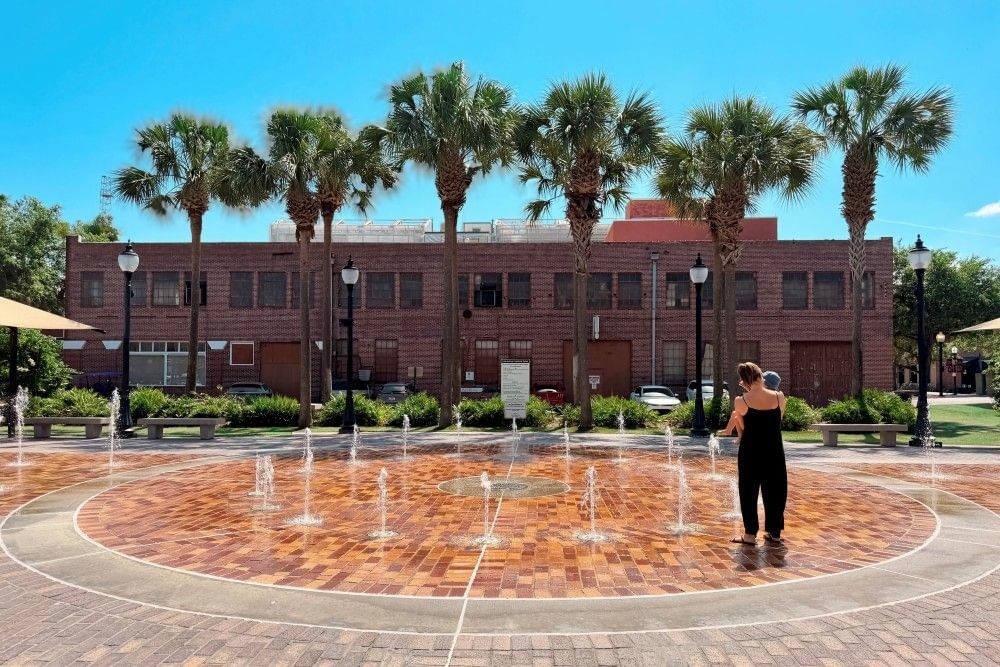 A cobblestone splash pad in front of a tall brick building, with a woman in a jumpsuit holding a baby facing away from the camera. 