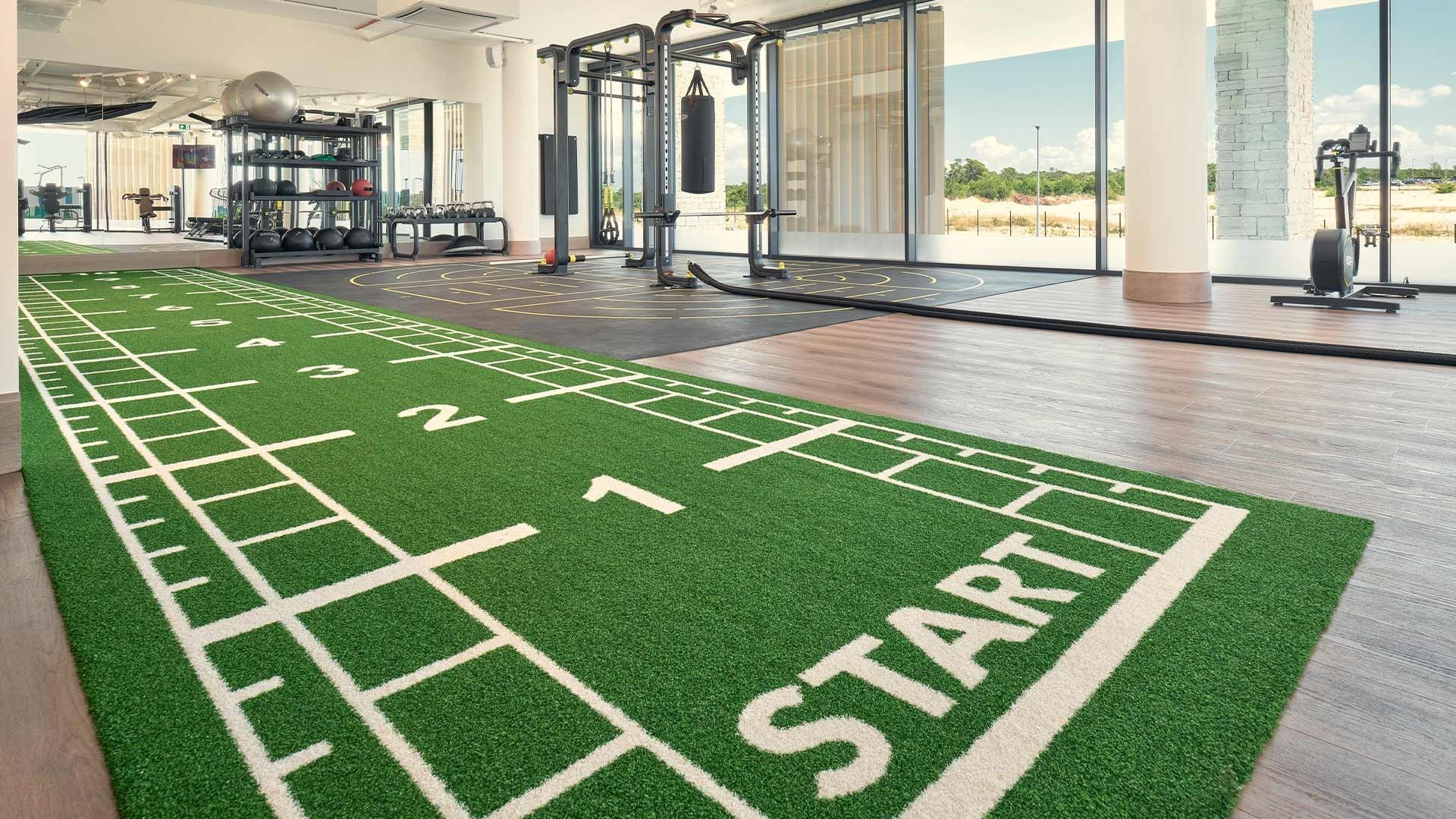 Fitnessstudio mit Startlinie auf grünem Teppich und verschiedenen Trainingsgeräten.