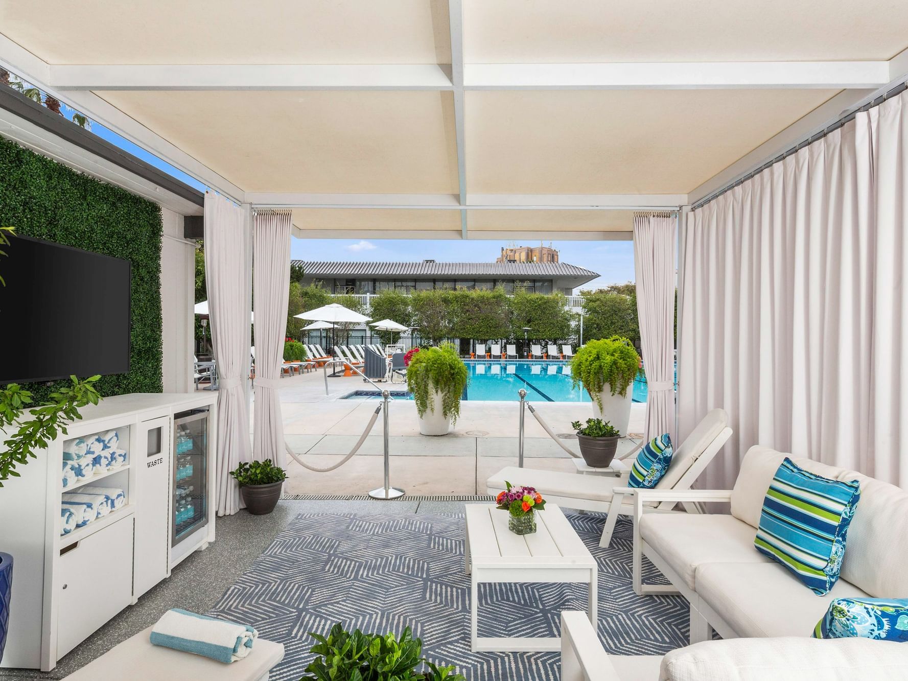 private cabana next to pool in background, cabana has white furniture, rug, small table, and wall-hung TV