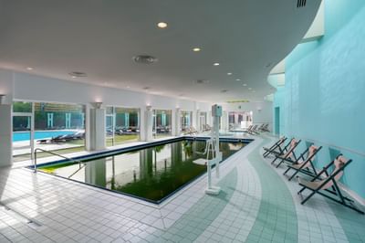 Indoor pool with greenish water and tiled flooring, surrounded by lounge chairs and large windows at Ana Hotels Europa