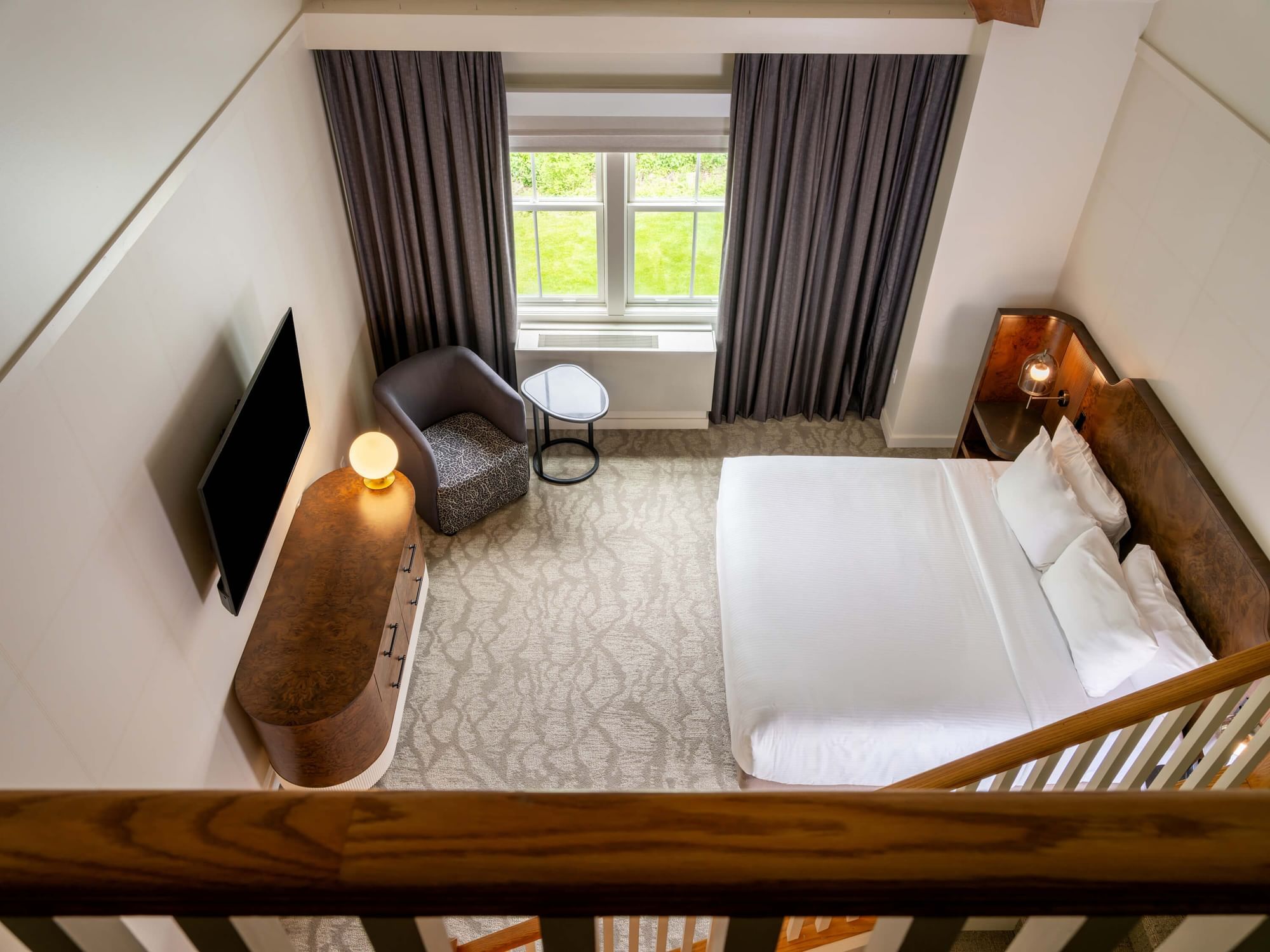 Top view of a lofted hotel room with a bed, armchair, table, TV, and window with curtains.