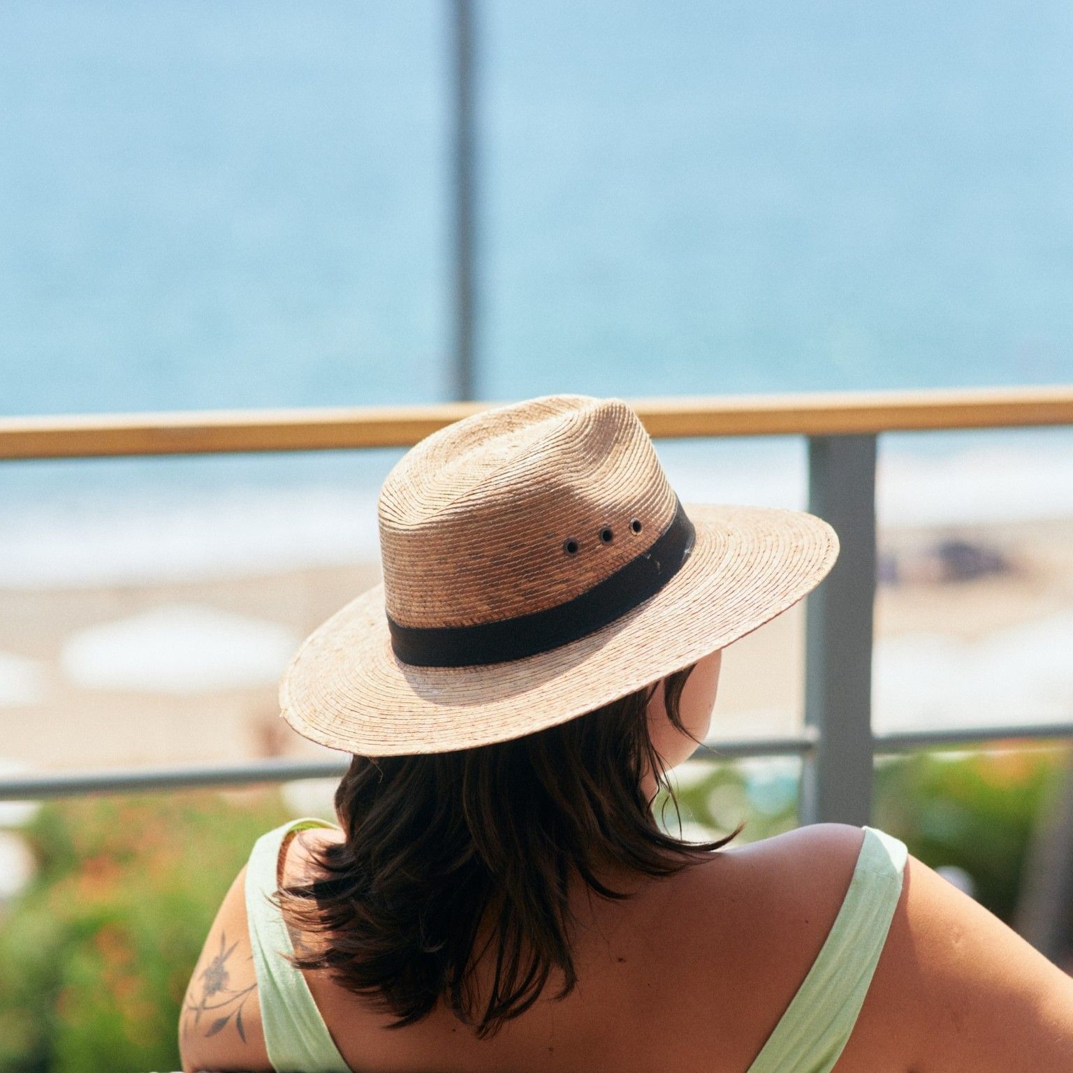 Woman in a green dress and straw hat overlooking the beach from a balcony.