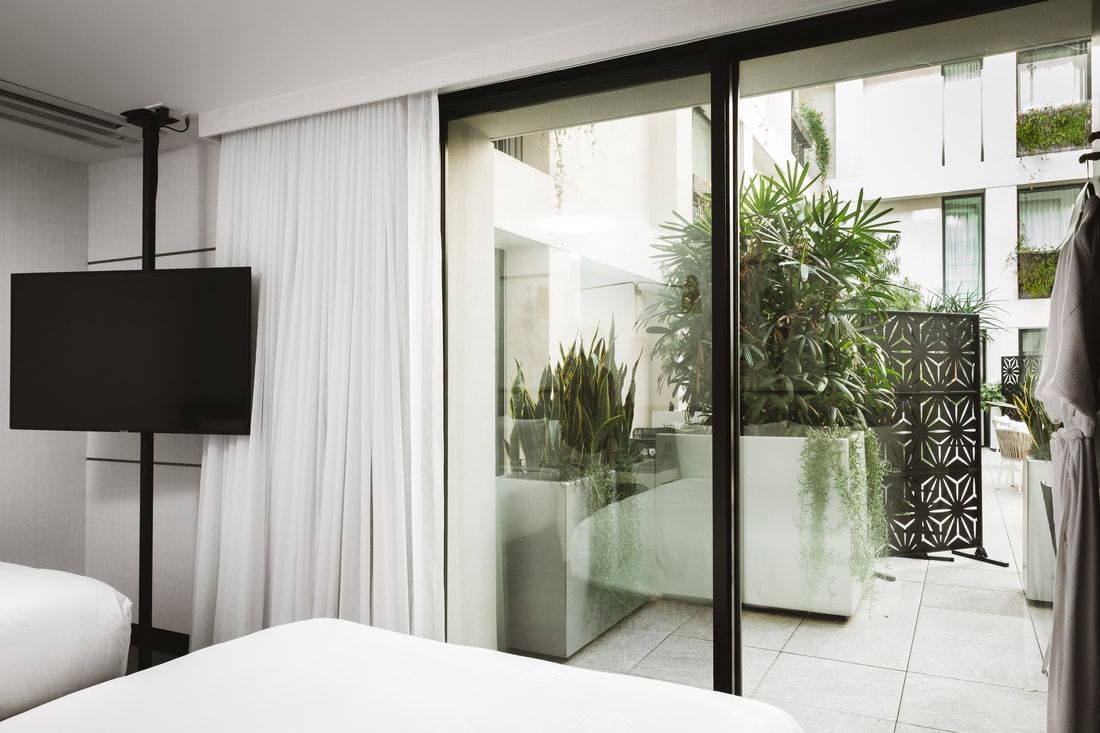 A hotel room with a bed, TV, and sliding glass door leading to a balcony with plants.