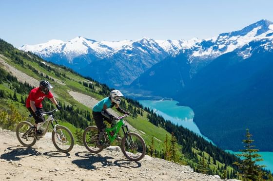 Two cyclists ride down a dirt trail with a view of a lake and snowy mountains at Aava Whistler.