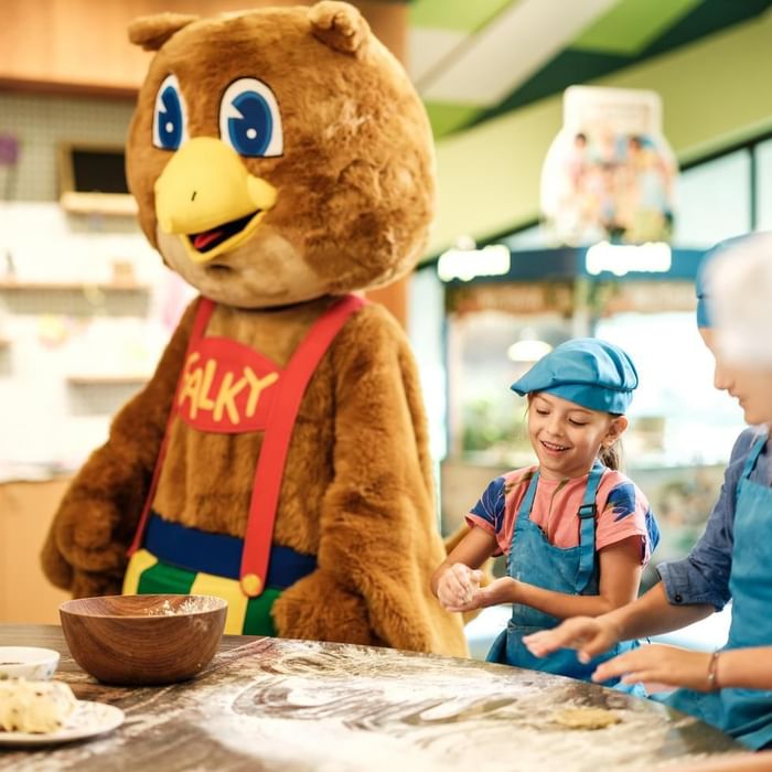 Two children baking cookies with a large brown mascot named Falky at a table.